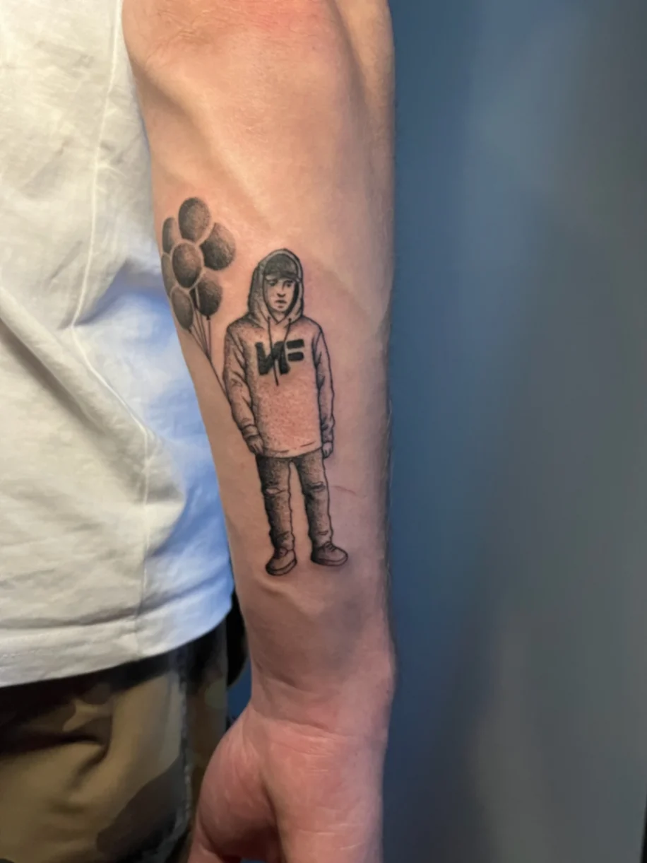 Black and grey figure in hoodie holding balloons tattooed on forearm
