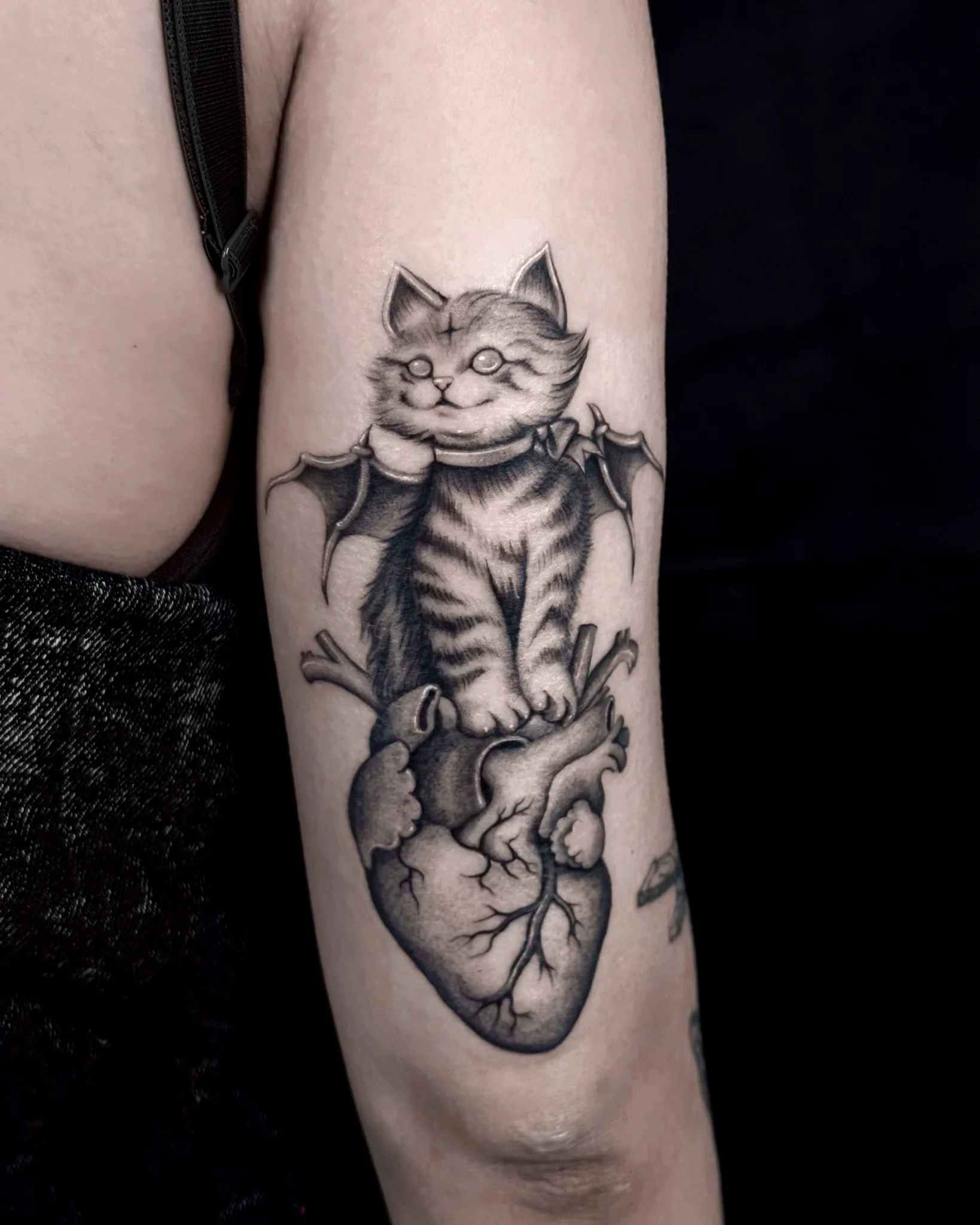 Black and grey demon kitten with bat wings clutching anatomical heart on upper arm