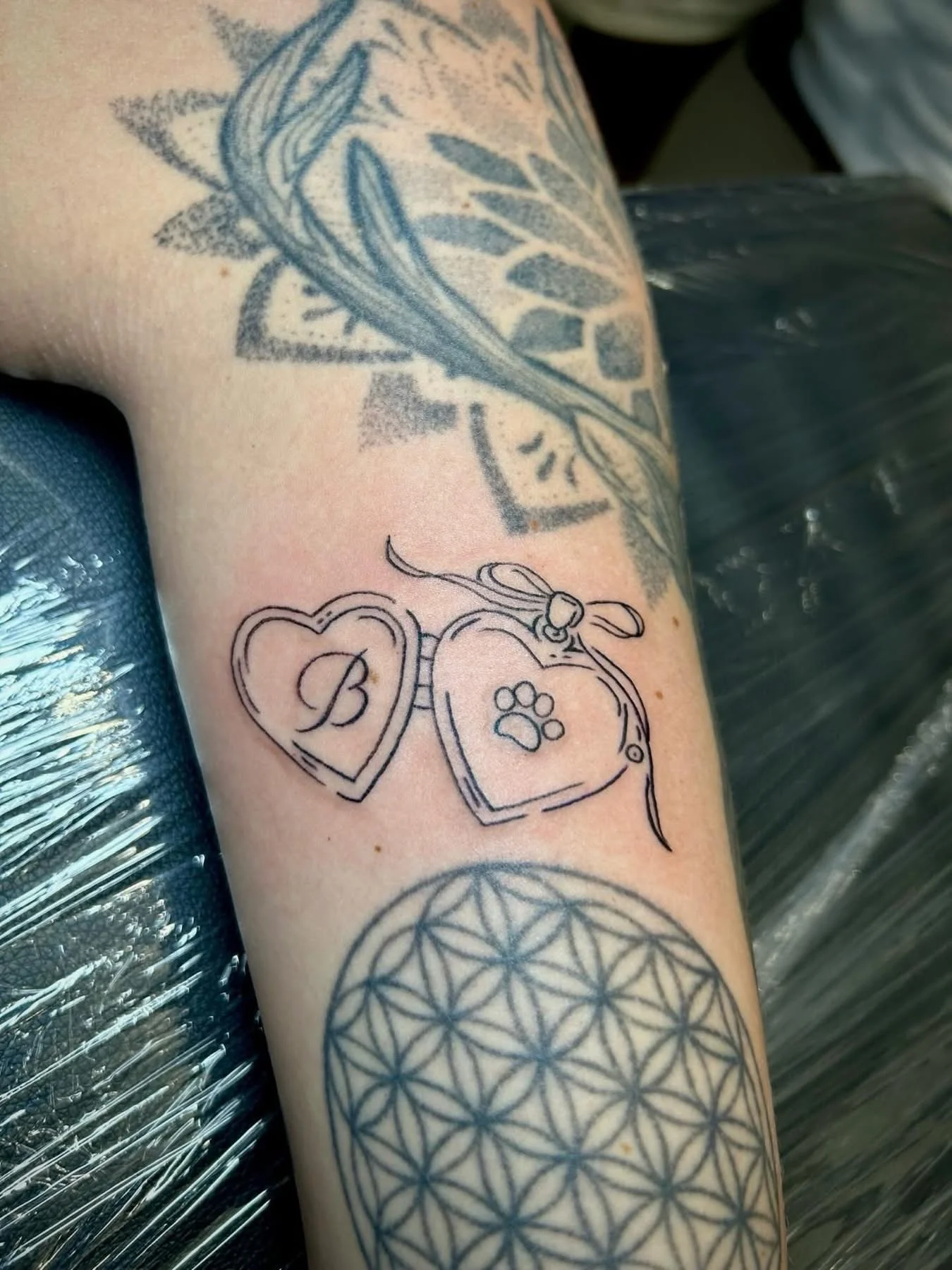 Fine line open heart locket tattoo with initial and paw print on forearm
