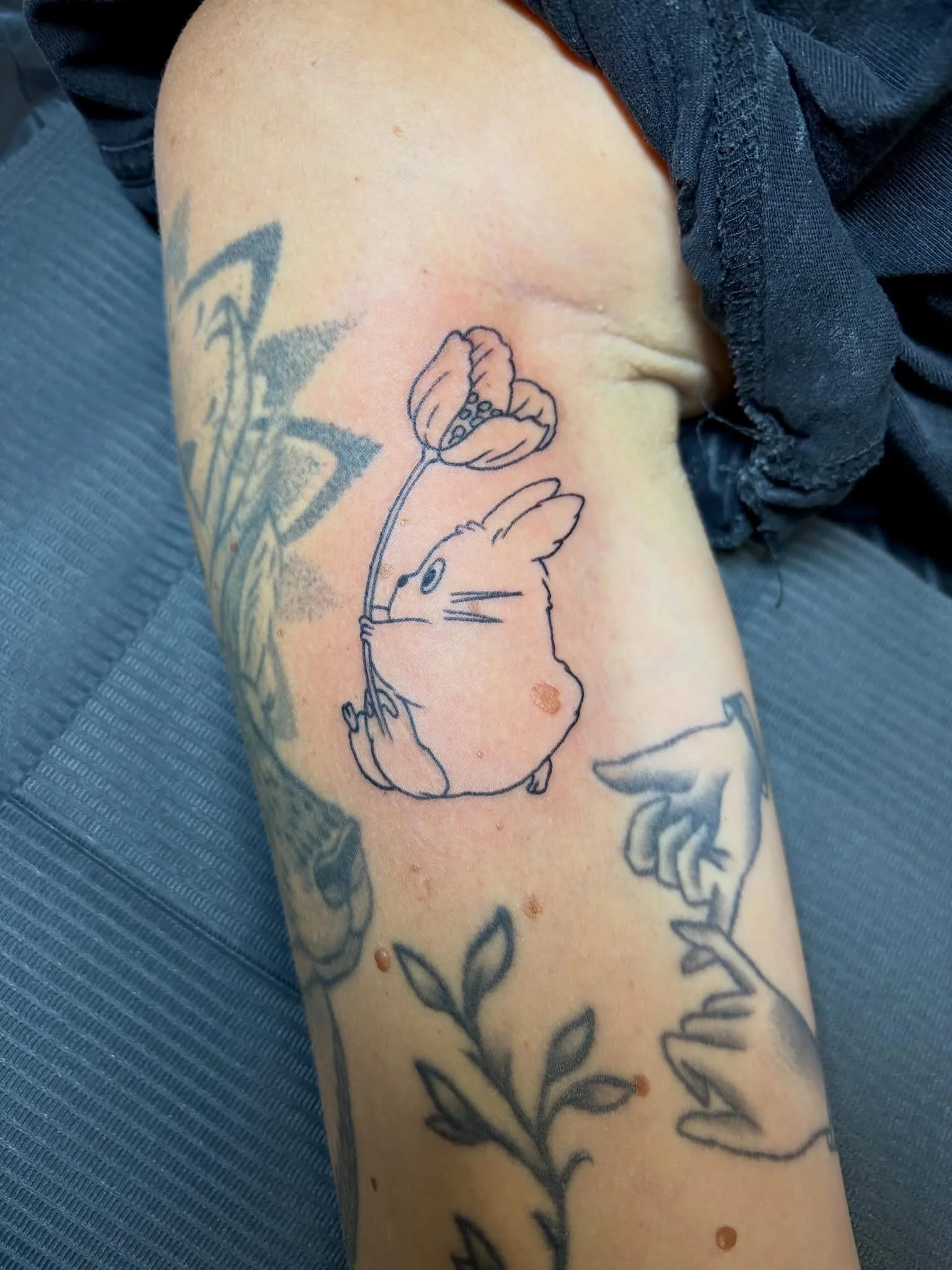 Fine line Totoro holding a flower tattoo on forearm