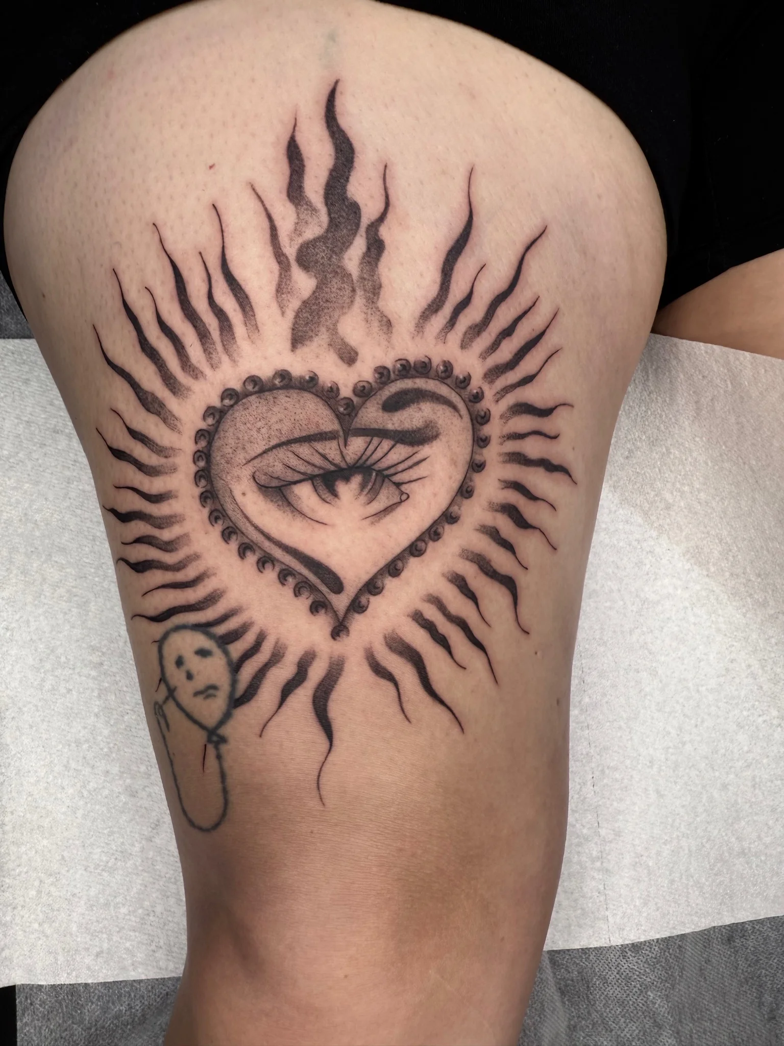 Black and grey sacred heart eye tattoo with rays on thigh