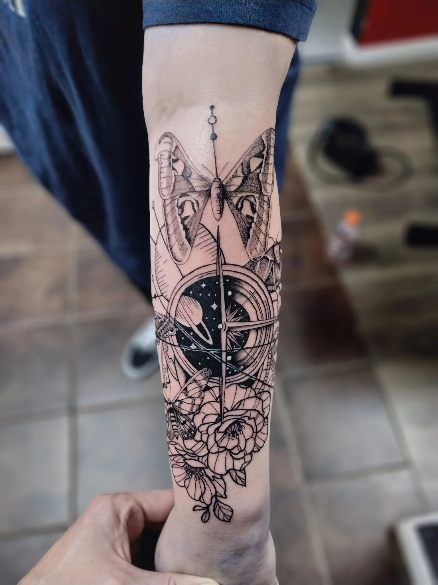 Illustrative blackwork moth compass and floral tattoo on forearm
