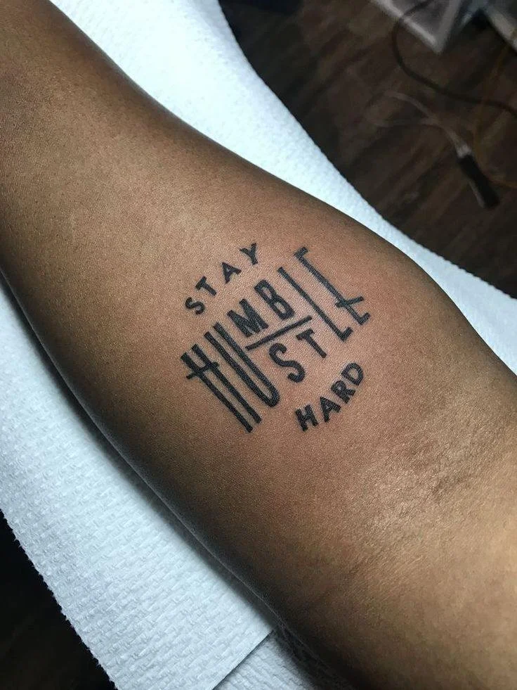 Bold typographic 'Stay Humble Hustle Hard' lettering tattoo on forearm